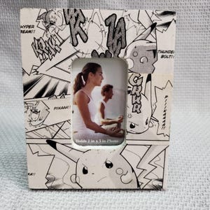 May include: A rectangular photo frame with a comic book design featuring Pikachu. The frame holds a 2 x 3 inch photo. The frame has black and white comic book panels with text like "HYPER BEAM!!" and "PIIKAAA!!".