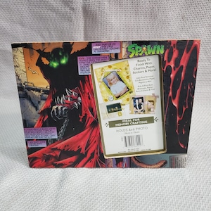 May include: A rectangular photo frame featuring comic book art of Spawn. The frame holds a 4x6 inch photo and includes text that reads "Ready To Finish With Charms, Paper, Stickers & More!" and "Ideal for Memory Crafting."