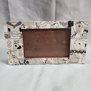 May include: Rectangular picture frame with a comic strip design featuring black and white illustrations of cartoon characters. The frame has a brown interior and is made of a light-colored material.