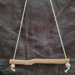 May include: A light-colored wooden rod suspended by a single piece of off-white rope. The rod features a natural, slightly irregular grain and texture. At each end of the rod, a small, smooth, round wooden bead is threaded onto the rope.