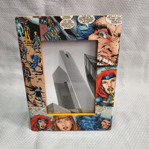 May include: A rectangular photo frame featuring comic book art. The frame is decorated with panels from a comic book, showcasing various characters and speech bubbles. The frame surrounds a photo of a modern building.