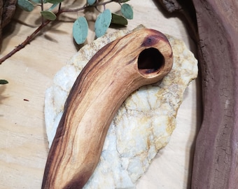 May include: A handcrafted wooden pipe with a curved design and a dark brown finish. The pipe features a circular opening at one end and a pointed tip. The wood grain is visible, showcasing natural patterns. The pipe is displayed on a light-colored stone.