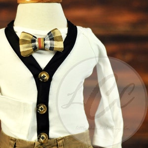 May include: A white long-sleeved onesie with a black trim and buttons. The onesie has a plaid bow tie with a red stripe.
