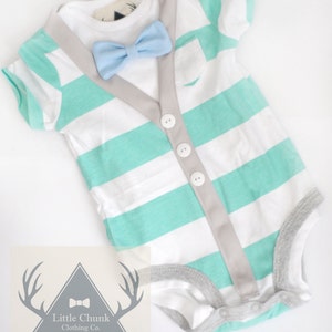 Può includere: Un body per neonato a righe bianche e verde menta con bordino grigio e un papillon azzurro chiaro. Il body ha una finta abbottonatura sul davanti e una piccola tasca sul petto. Il body è etichettato "Little Chunk Clothing Co. Est. 2013."