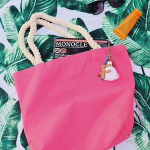 May include: A pink canvas tote bag with a rope handle. The bag is sitting on a white and green patterned beach towel. A magazine titled "Monocle" is partially visible inside the bag.