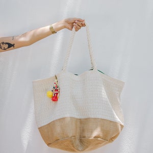 Puede incluir: Una bolsa de tela blanca y marr&oacute;n con asas de cuerda. La bolsa tiene una parte superior blanca tejida y una parte inferior marr&oacute;n. Un borla de colores cuelga de la bolsa.