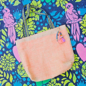 May include: A light pink tote bag with a woven texture and colorful straps. The bag has a tassel charm with colorful beads and a pink and blue fringe. The bag is on a dark blue background with a floral pattern.