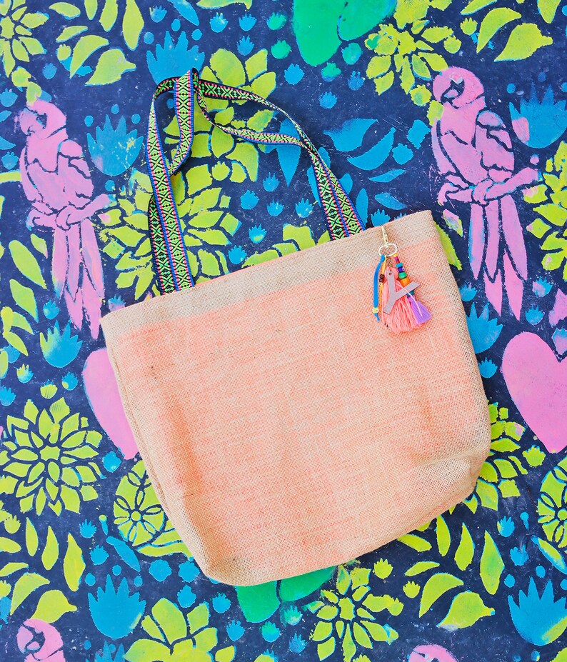 May include: A light pink tote bag with a woven texture and colorful straps. The bag has a tassel charm with colorful beads and a pink and blue fringe. The bag is on a dark blue background with a floral pattern.