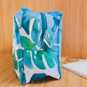 May include: A blue and green toiletry bag with a zipper closure and a white background. The bag features a watercolor-style print of tropical leaves.