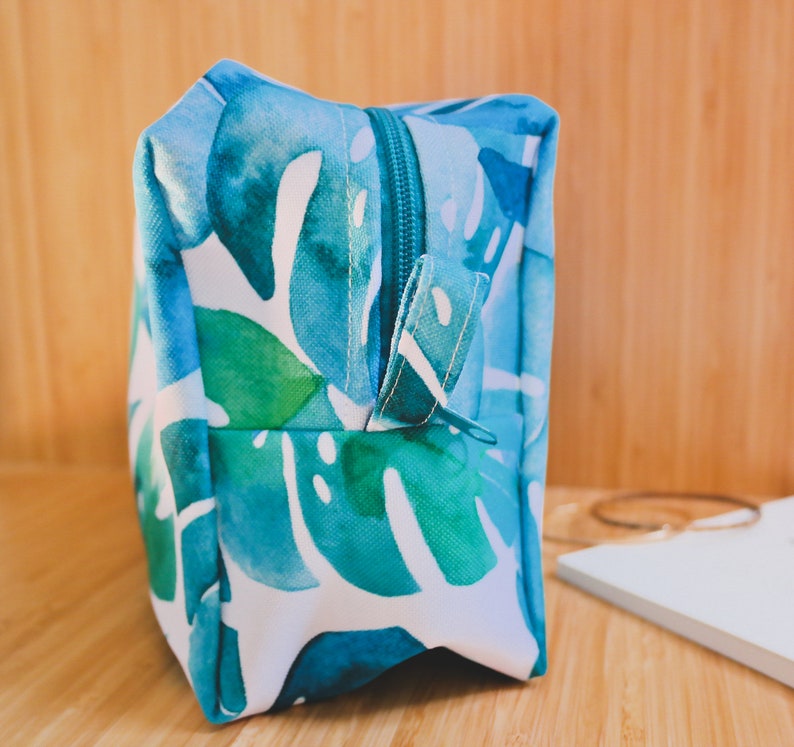 May include: A blue and green toiletry bag with a zipper closure and a white background. The bag features a watercolor-style print of tropical leaves.