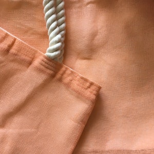 May include: A close-up of a peach-coloured mesh tote bag with a white rope handle. The bag is made of a soft, lightweight material and has a simple, classic design.