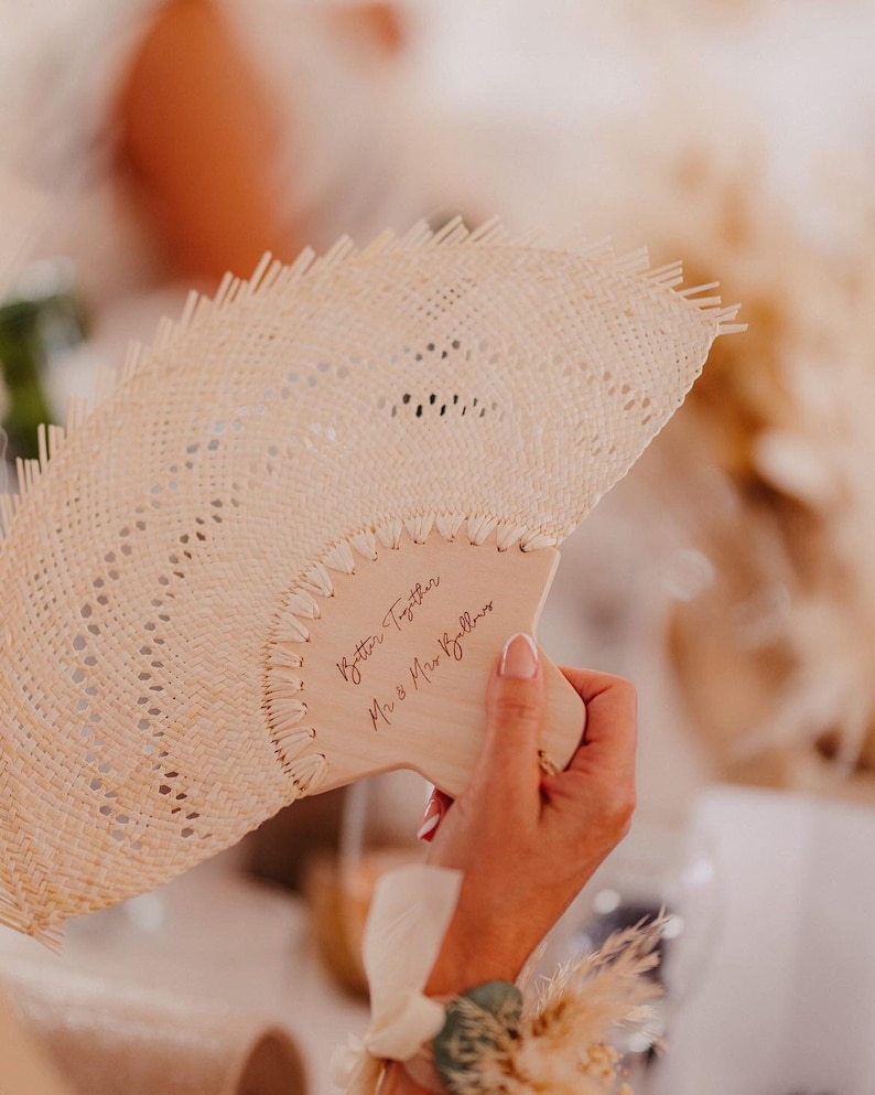 A hand holding a woven palm fan with "Better Together" engraved on the wooden handle.