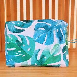 May include: A white zippered pouch with a tropical leaf print in shades of green and blue. The pouch has a white zipper and a small loop on the side.