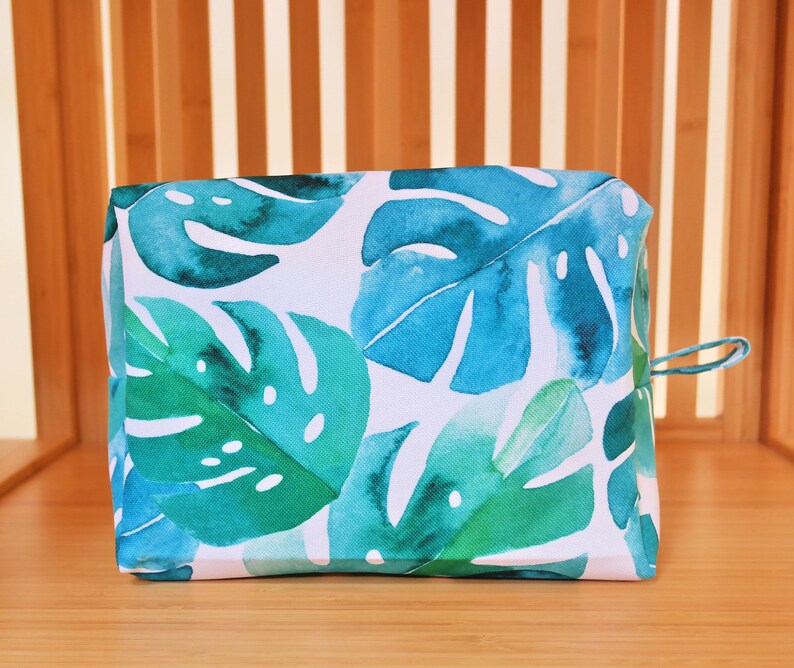 May include: A white zippered pouch with a tropical leaf print in shades of green and blue. The pouch has a white zipper and a small loop on the side.