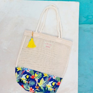 May include: A white mesh tote bag with a blue and green tropical print on the bottom. The bag has a yellow tassel attached to the handle and the text "LA BOHEME" is printed on the front.