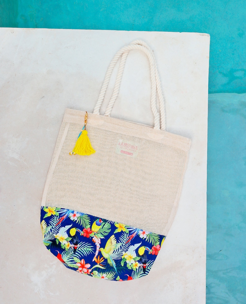 May include: A white mesh tote bag with a blue and green tropical print on the bottom. The bag has a yellow tassel attached to the handle and the text "LA BOHEME" is printed on the front.