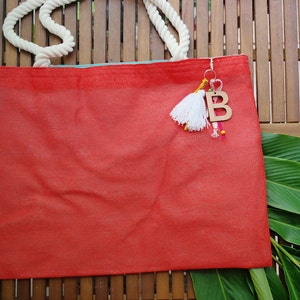 May include: A red tote bag with white rope handles and a wooden letter "B" charm with colourful tassels hanging from it.
