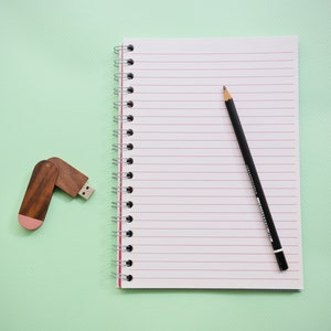 Può includere: Un quaderno bianco con carta rigata rossa e una rilegatura a spirale argentata. Una matita nera è appoggiata sul quaderno. Un'unità flash USB marrone e rosa si trova sul lato sinistro dell'immagine.