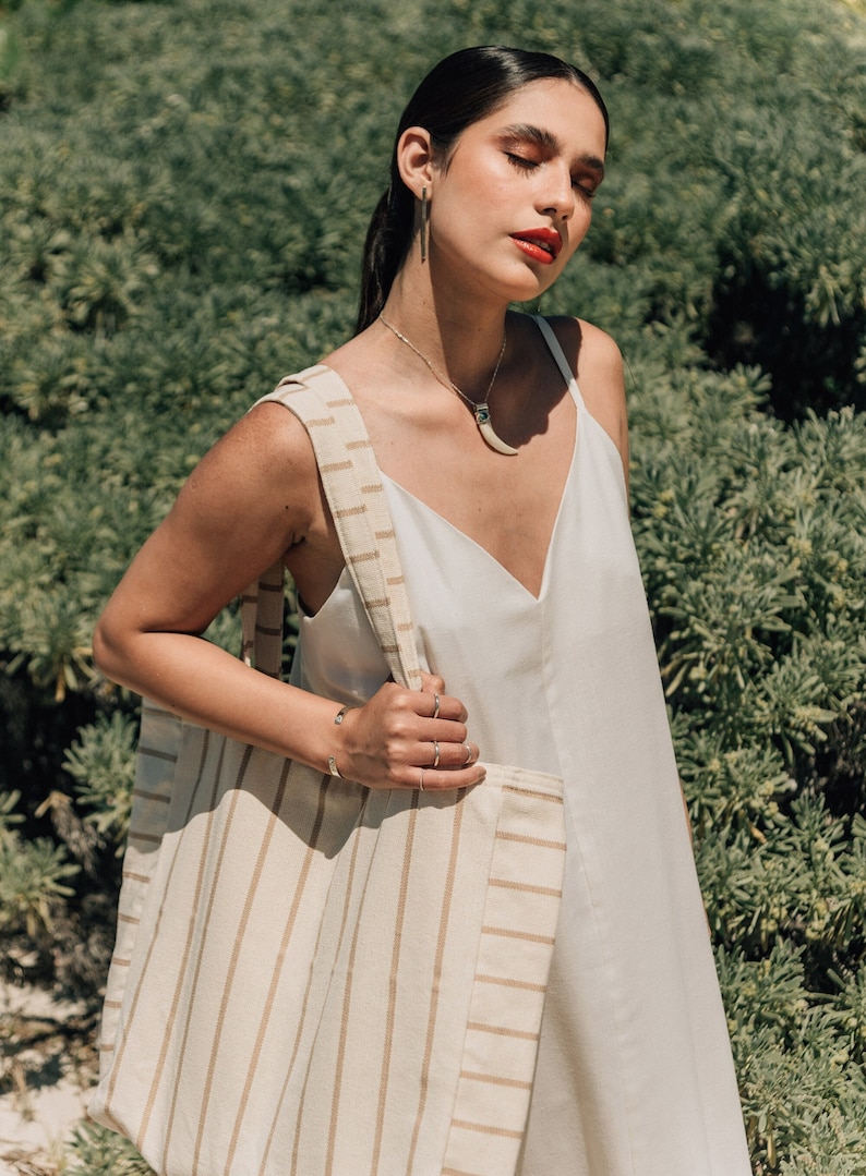 May include: A woman wearing a white sleeveless dress with a brown and white striped tote bag.