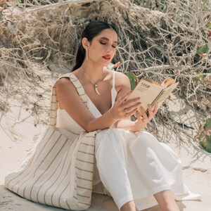 May include: A woman wearing a white dress and sandals sits on a sandy beach, reading a book. She is carrying a striped tote bag.