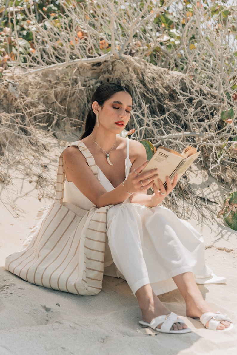 May include: A woman wearing a white dress and sandals sits on a sandy beach, reading a book. She is carrying a striped tote bag.