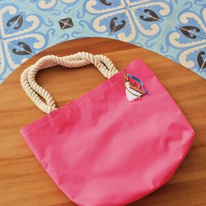 May include: A pink canvas tote bag with a white rope handle and a charm attached to the handle. The charm features a white tassel and a wooden letter 'F'.