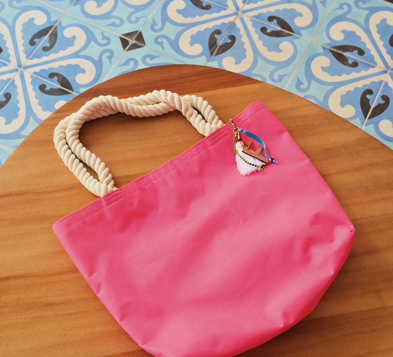 May include: A pink canvas tote bag with a white rope handle and a charm attached to the handle. The charm features a white tassel and a wooden letter 'F'.