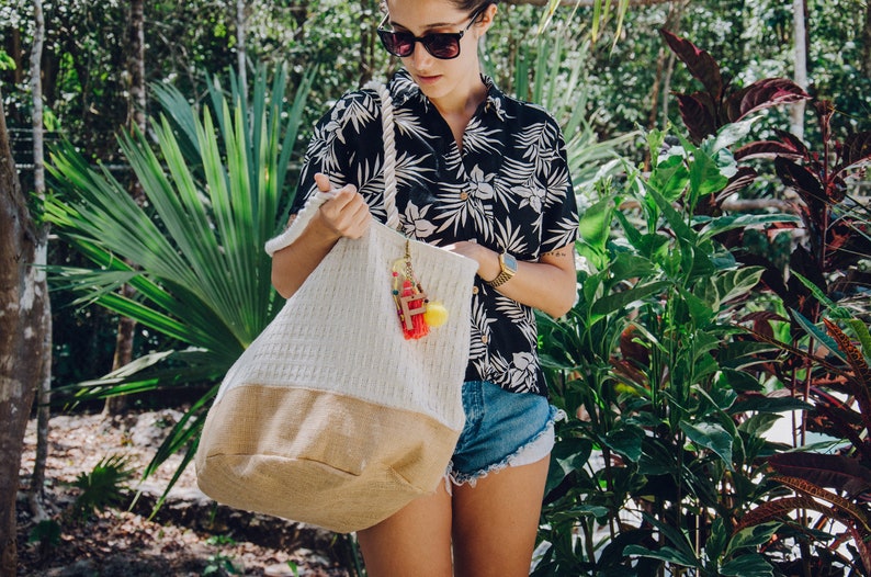 Puede incluir: Una mujer con una camisa de flores blancas y negras y pantalones cortos de mezclilla est&aacute; sosteniendo una bolsa de mano blanca y tostada con un asa de cuerda. La bolsa tiene un peque&ntilde;o amuleto adjunto.