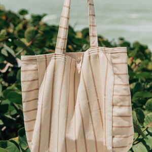 May include: A large, striped tote bag made of natural fabric. The bag has brown stripes on a cream background. The bag is being held by a hand.