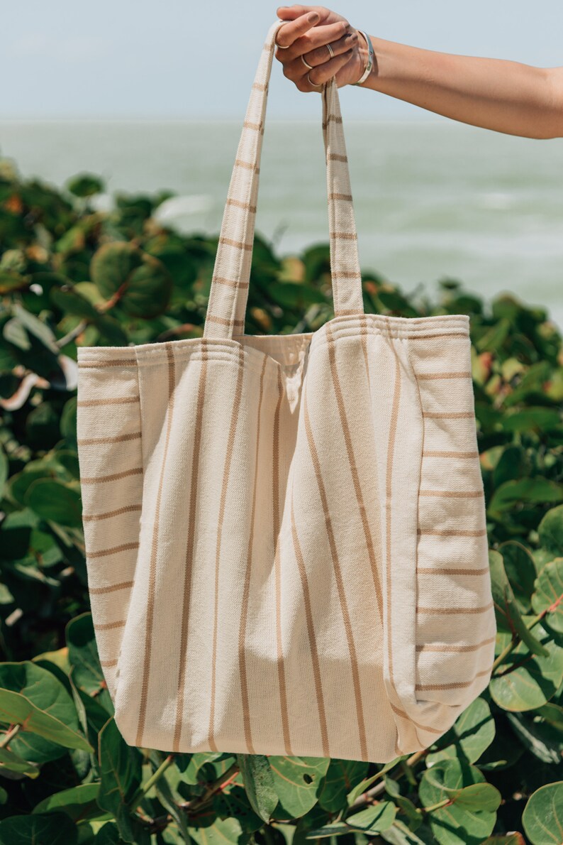 May include: A large, striped tote bag made of natural fabric. The bag has brown stripes on a cream background. The bag is being held by a hand.
