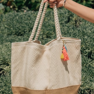 Puede incluir: Una bolsa de mano beige y marr&oacute;n con asas de cuerda y un abalorio de borla de colores. La bolsa tiene un patr&oacute;n de chevron y un fondo marr&oacute;n s&oacute;lido.