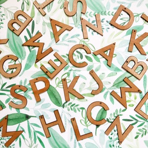 May include: Wooden letters of the alphabet, painted in a light brown color, are scattered on a white background with a green watercolor leaf pattern.