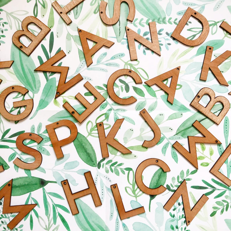 May include: Wooden letters of the alphabet, painted in a light brown color, are scattered on a white background with a green watercolor leaf pattern.