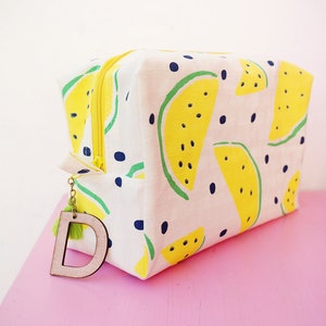 Puede incluir: Una bolsa blanca con cremallera con estampado de sandía amarilla y lunares negros. La bolsa tiene una cremallera amarilla y un dije de madera con la letra "D".