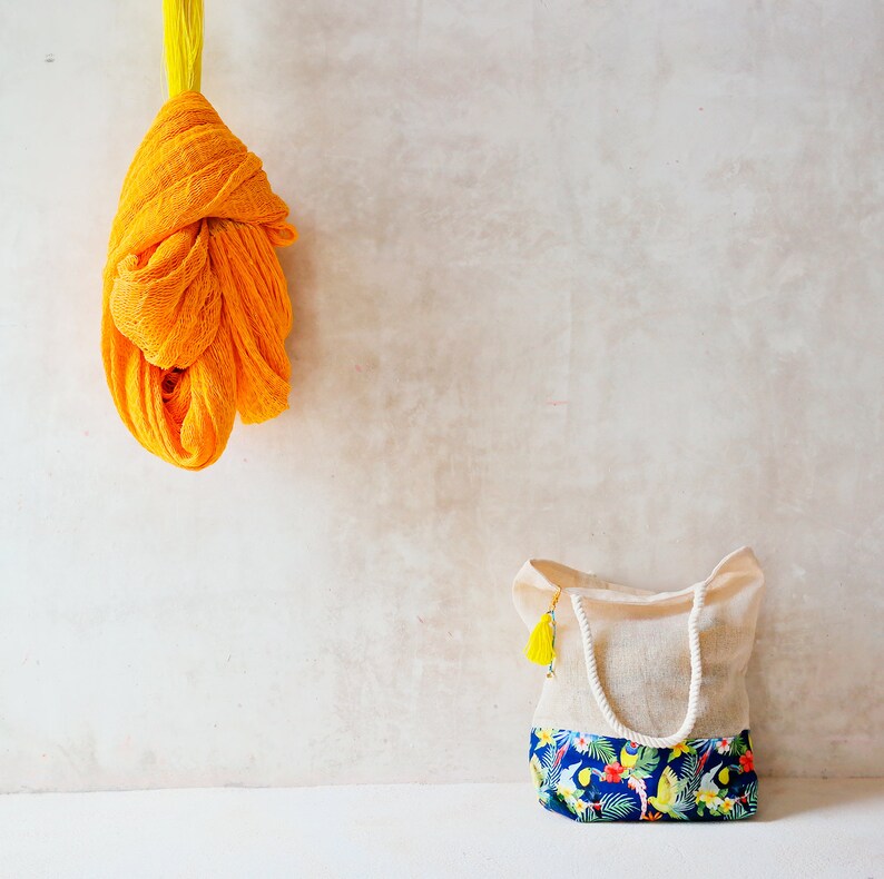 May include: A yellow crocheted fabric hanging on a wall next to a white tote bag with a floral print. The tote bag has a yellow tassel.
