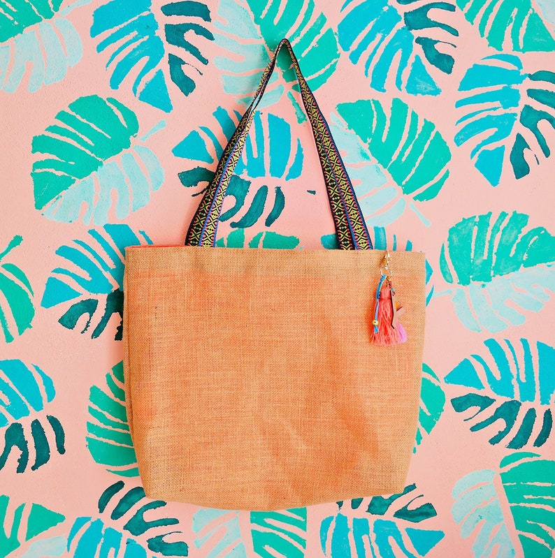 May include: A light orange tote bag with a woven texture and black and blue patterned straps. The bag is hanging against a pink background with a blue and green tropical leaf pattern.
