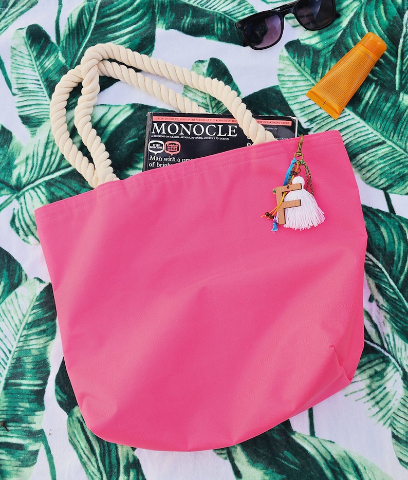 May include: A pink canvas tote bag with a rope handle. The bag is sitting on a white and green patterned beach towel. A magazine titled "Monocle" is partially visible inside the bag.