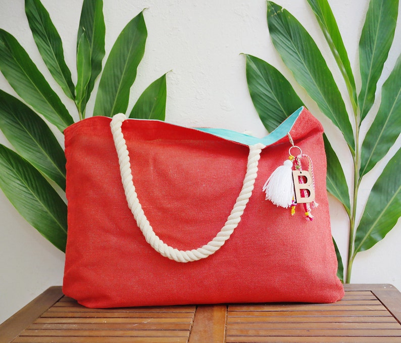 May include: A red canvas tote bag with white rope handles and a turquoise lining. The bag has a wooden letter "B" charm attached to it.