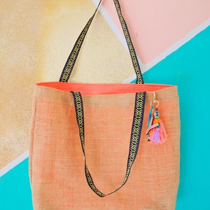 May include: A large, peach-colored tote bag made of woven fabric with black and yellow patterned straps. The bag has a small, colorful tassel hanging from the handle.