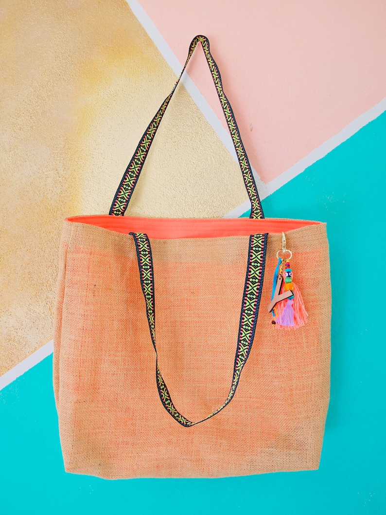 May include: A large, peach-colored tote bag made of woven fabric with black and yellow patterned straps. The bag has a small, colorful tassel hanging from the handle.