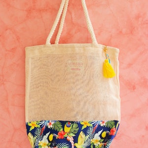 May include: A beige tote bag with a blue and green tropical print on the bottom. The bag has a rope handle and a yellow tassel.