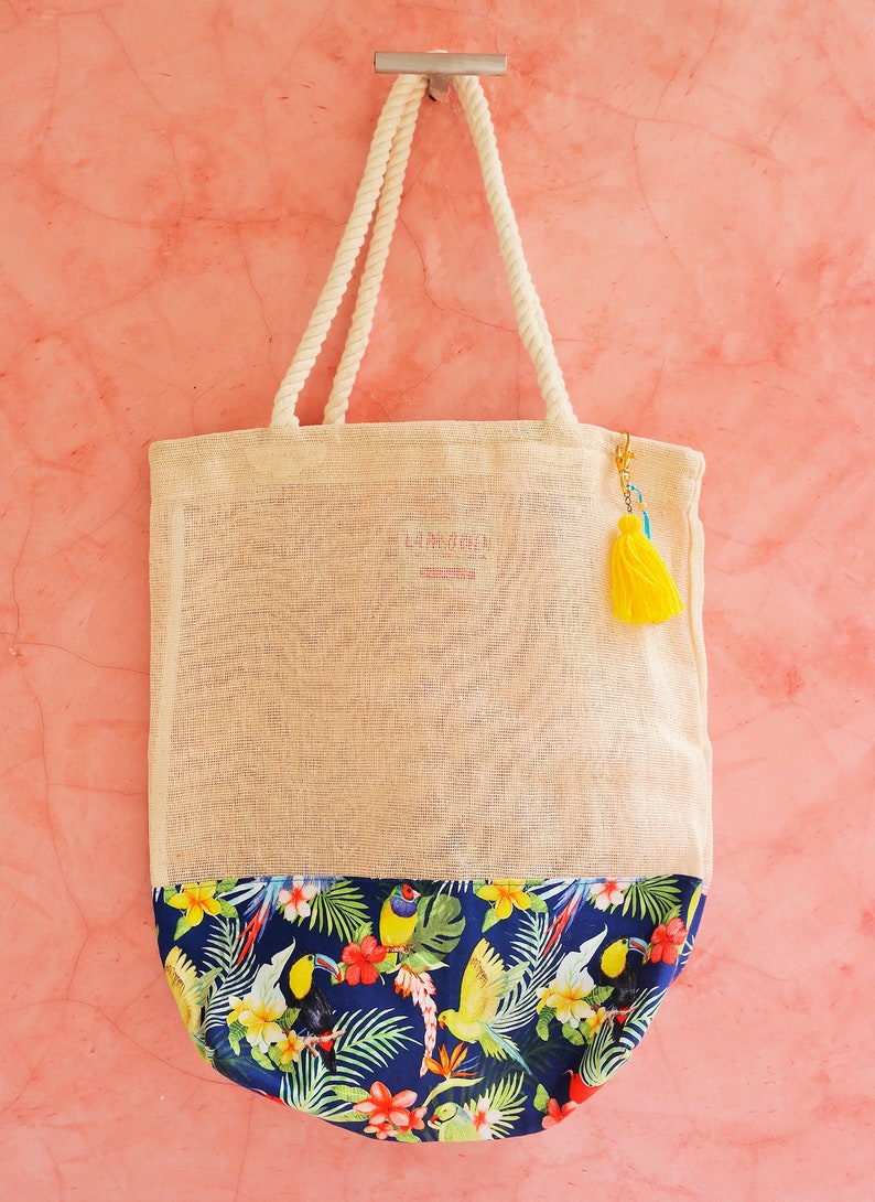 May include: A beige tote bag with a blue and green tropical print on the bottom. The bag has a rope handle and a yellow tassel.