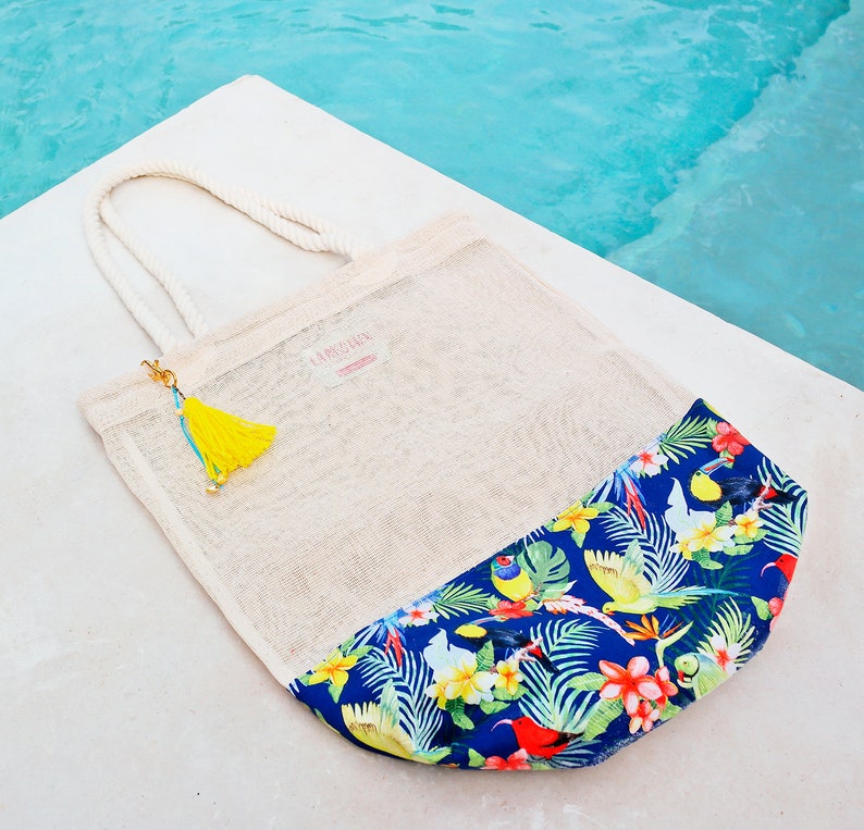 May include: A white mesh tote bag with a tropical print on the bottom half. The bag has a yellow tassel and the text "La Boheme" on the front.