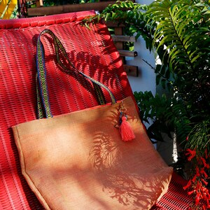 May include: A tan jute tote bag with a colorful woven strap and a pink tassel charm. The bag is sitting on a red wicker chair.