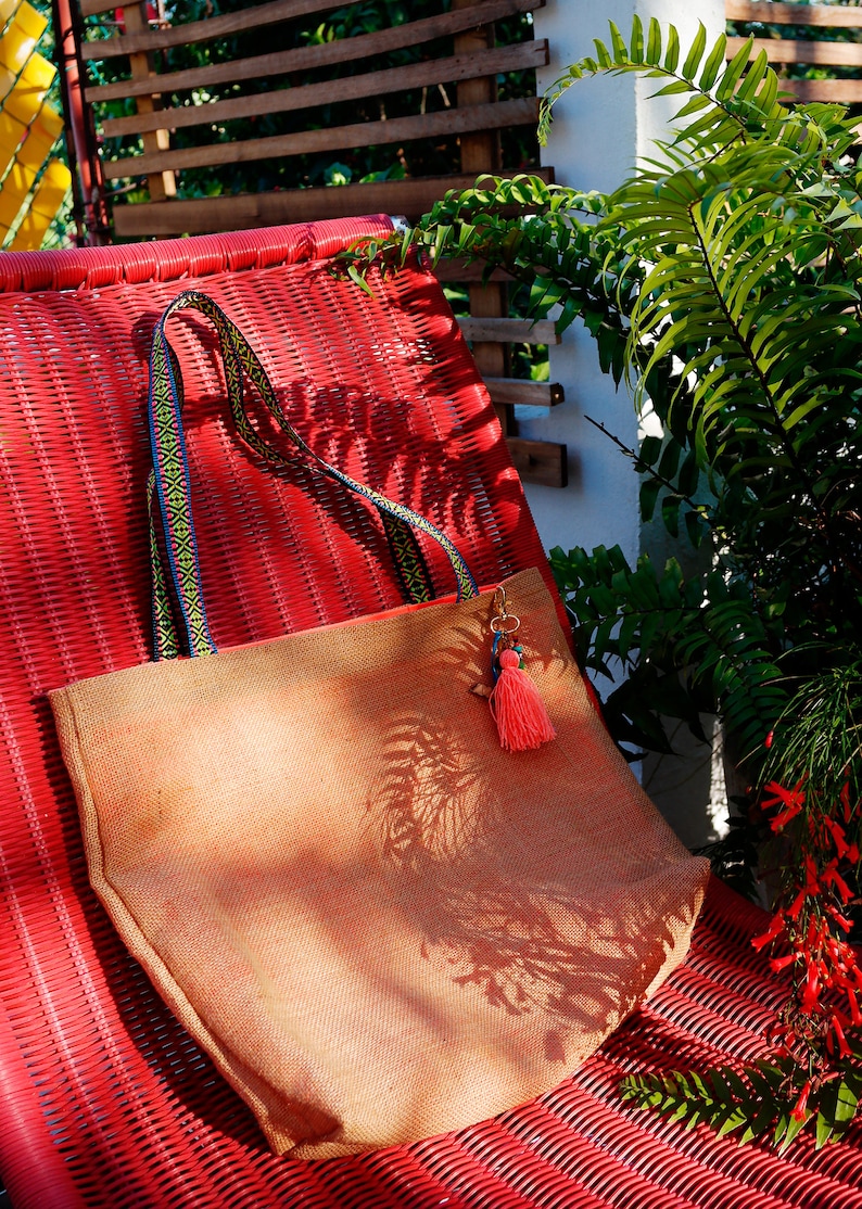 May include: A tan jute tote bag with a colorful woven strap and a pink tassel charm. The bag is sitting on a red wicker chair.