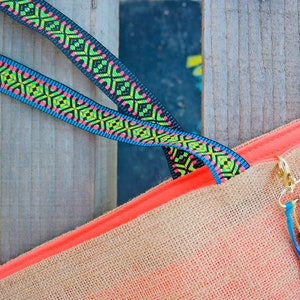 May include: A close-up of a jute tote bag with a bright orange trim and two black and multi-colored straps. The straps have a geometric pattern.