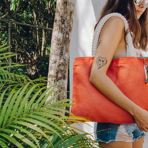 May include: A woman is carrying a large, bright orange tote bag with a white rope handle and a green and blue tassel. The bag is made of canvas and has a heart-shaped tattoo on the woman's arm.