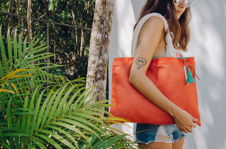 May include: A woman is carrying a large, bright orange tote bag with a white rope handle and a green and blue tassel. The bag is made of canvas and has a heart-shaped tattoo on the woman's arm.