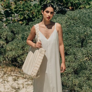 May include: A woman wearing a white maxi dress with a ruffled hem and a striped tote bag. She is standing in front of a green bush.
