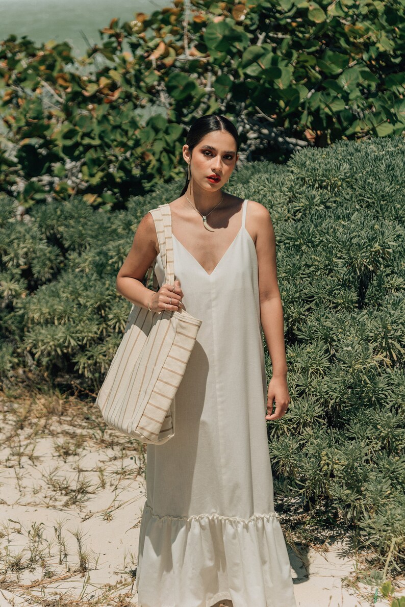 May include: A woman wearing a white maxi dress with a ruffled hem and a striped tote bag. She is standing in front of a green bush.